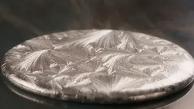 Molten tin being poured onto a cold glass surface freezing into a branching dendritic fern pattern, camera arcing countercloc