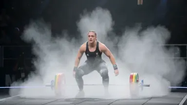 Weightlifter dropping a loaded barbell behind her head in a failed snatch attempt on a competition platform, camera on a slow