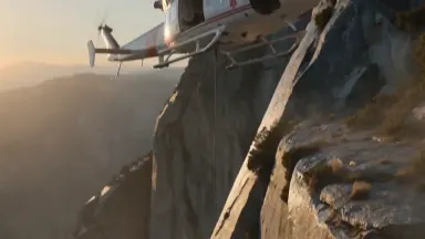 Rescue helicopter hovering beside a sheer granite cliff face as a stretcher is winched steadily upward on a cable, camera cra
