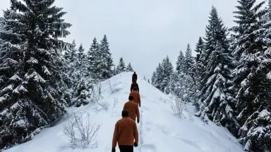 The models continue walking steadily single-file along the ridge away from camera. Camera tilts slowly upward from the snow-p