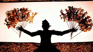 Crane the camera slowly upward from the lamp and batik border past the puppeteer's silhouette to the top of the kelir screen,