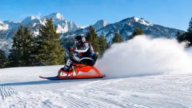 The sit-skier drives through the arc and the spray plume explodes outward then settles, the skier transitions immediately int