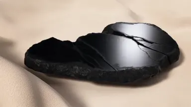 Freshly split obsidian resting on pale wrinkled linen, the conchoidal fracture surface a deep black mirror catching a single 