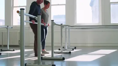 Physical therapist guiding a stroke patient through first standing steps between parallel bars in a bright rehabilitation gym