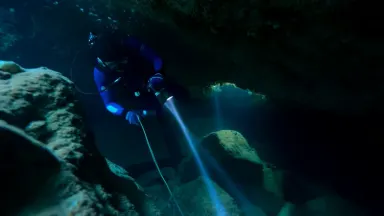 Clockwise orbit around a cave diver reeling a bright nylon guide line through a collapsed passage in a dark limestone cavern,
