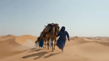 Fully loaded freight camel caravan crests a Mauritanian dune ridge at midday, handlers walking alongside in loose robes, slow