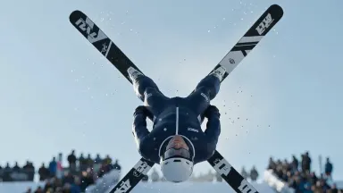 Freestyle ski aerialist rotating fully inverted above a kicker ramp, skis crossed at the apex of a layout, slow push in tight