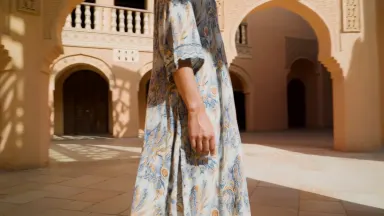 A model in a floor-length hand-painted batik robe standing motionless inside a sunlit Moroccan courtyard, a single gust of wi