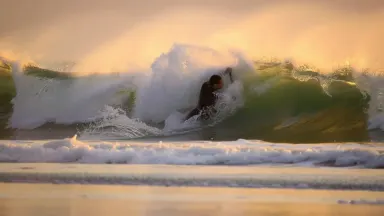 Competitive paddleboarder buried to the waist in a collapsing shore break wave on a black sand beach, white water engulfing t