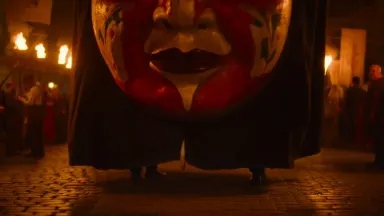 Giant carnival head puppet bobbing through a cobblestone festival square, papier-mache face painted in vivid vermillion and g