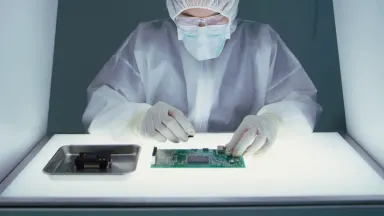 Lab technician in a clean room suit assembling a circuit board for a medical imaging device beneath a particle-free laminar f