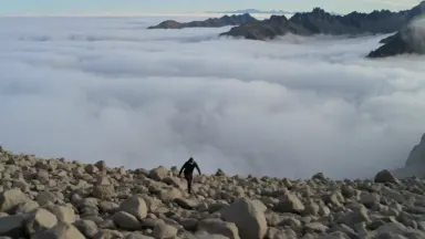 Mountain ultrarunner in technical gear scrambling across a chaotic field of loose scree boulders above a solid cloud layer, b