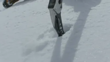 Ice axe spike biting into steep hard snow mid self-arrest, loose spindrift exploding outward from the arrest point and stream