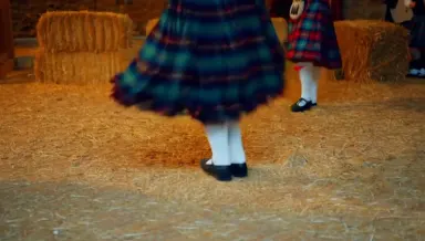 Ceilidh dancers in tartan spin mid-reel inside a stone Highland barn strung with warm tungsten lights, their skirts and kilts