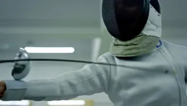 A competitive fencer in white jacket and wire mask lunges into full extension, blade tip striking the opponent's mask in a bu