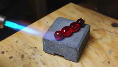 A goldsmith holds a handheld torch over a hollow-form brooch resting on a grey charcoal block, the metal warming from dull to