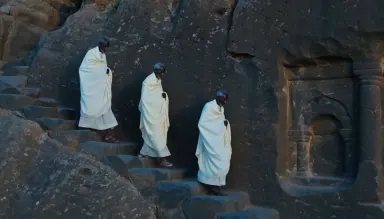 Ancient rock-hewn church carved directly into a basalt cliff face on an Ethiopian escarpment, priests descending exterior ste