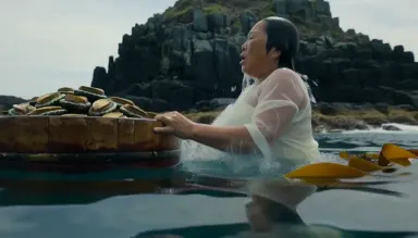 Traditional Japanese ama diver surfacing beside a wooden tub, abalone shells heaped inside, sea cliff rising behind. Camera o