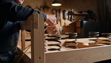 Upholsterer hand-tying a traditional eight-way spring coil into a bare hardwood sofa frame in a workshop, arc around the subj
