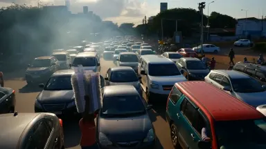 Lottery ticket vendor threading through a gridlocked intersection in Lagos at midday, balancing a tall stack of numbered slip