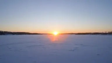 Sun dogs flanking a low winter sun over a frozen Finnish lake, all three lights glowing in a pale horizontal arc. The ice sur
