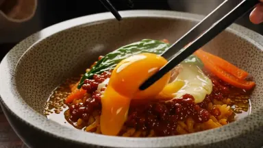 Overflowing bowl of bibimbap at the moment the raw egg yolk breaks as chopsticks begin to toss the ingredients, crane rising 