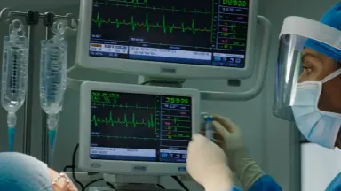 Anesthesiologist monitoring vital signs displays above a draped patient at the head of a surgical table during a long procedu