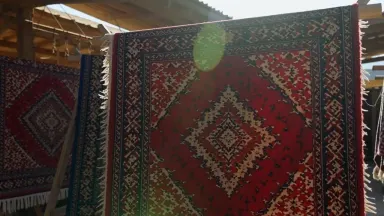 A collection of richly patterned pile rugs hangs draped over a heavy wooden rack in an open Anatolian dye market under harsh 