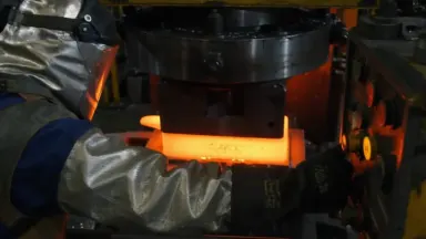A forge press operator in heat-shielded gear initiates a thousand-ton ram that descends onto a glowing orange titanium billet