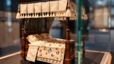 An exquisitely detailed 1:12 scale Elizabethan four-poster bed with hand-stitched linen canopy sits inside a glass display ca