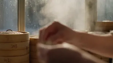 Dumpling maker pinching pleats into xiao long bao beside a window steamed opaque by a bank of bamboo steamers, static frame w