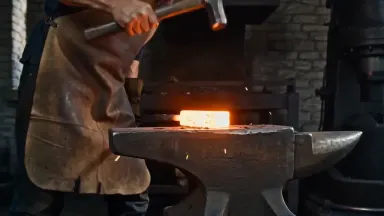 Knife forger folding high-carbon billet steel on an anvil with a cross-peen hammer mid-strike, tracking shot alongside at wai