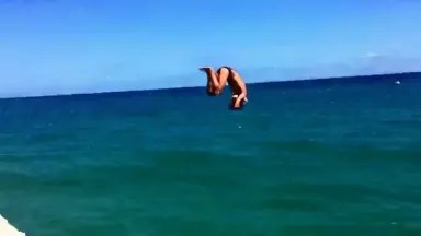A cliff diver launches from the edge of a Sardinian sea stack at midday and enters the turquoise water below in a perfect tig