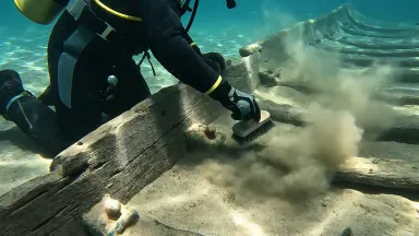 Underwater archaeologist in a dry suit brushing sediment from the curved ribs of a Bronze Age shipwreck hull resting on a pal