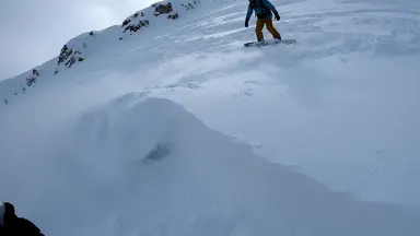 Backcountry snowboarder triggering a small controlled avalanche on a steep test slope, a white wave of powder spilling and sp