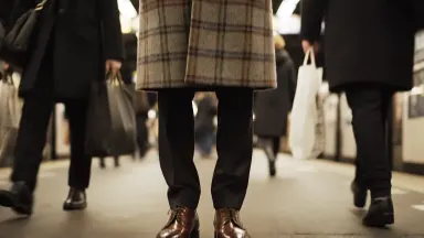 A model in a sharply tailored plaid overcoat standing perfectly still on a crowded London commuter platform, surrounding figu