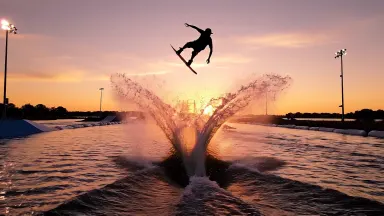 Competitive wakeboard rider launching a heelside backroll above a cable park lake at golden sunset, the board tail leaving a 