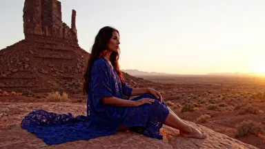 A model in a head-to-toe hand-embroidered indigo kaftan seated cross-legged atop a weathered desert mesa at golden hour, came