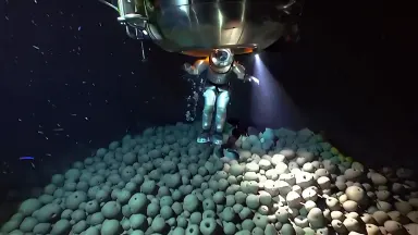 Saturation diver exiting a deep-sea bell into a black abyss above a manganese nodule field, crane move rising slowly from nod