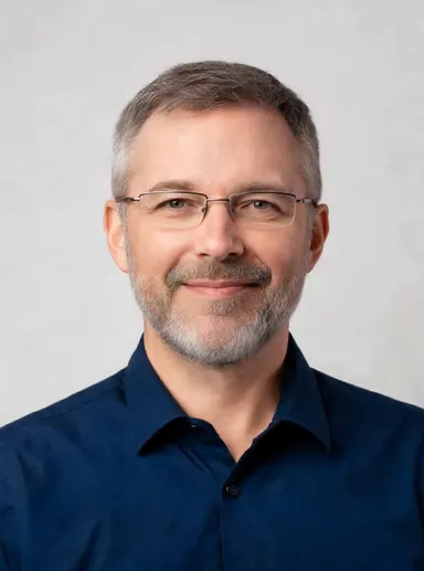 Studio portrait of a man in his 40s with short grey beard and glasses, navy shirt, confident warm expression, soft neutral ba