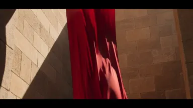 One continuous shot of a long bolt of crimson silk caught in a warm updraft inside a sunlit stone courtyard, the fabric unfur